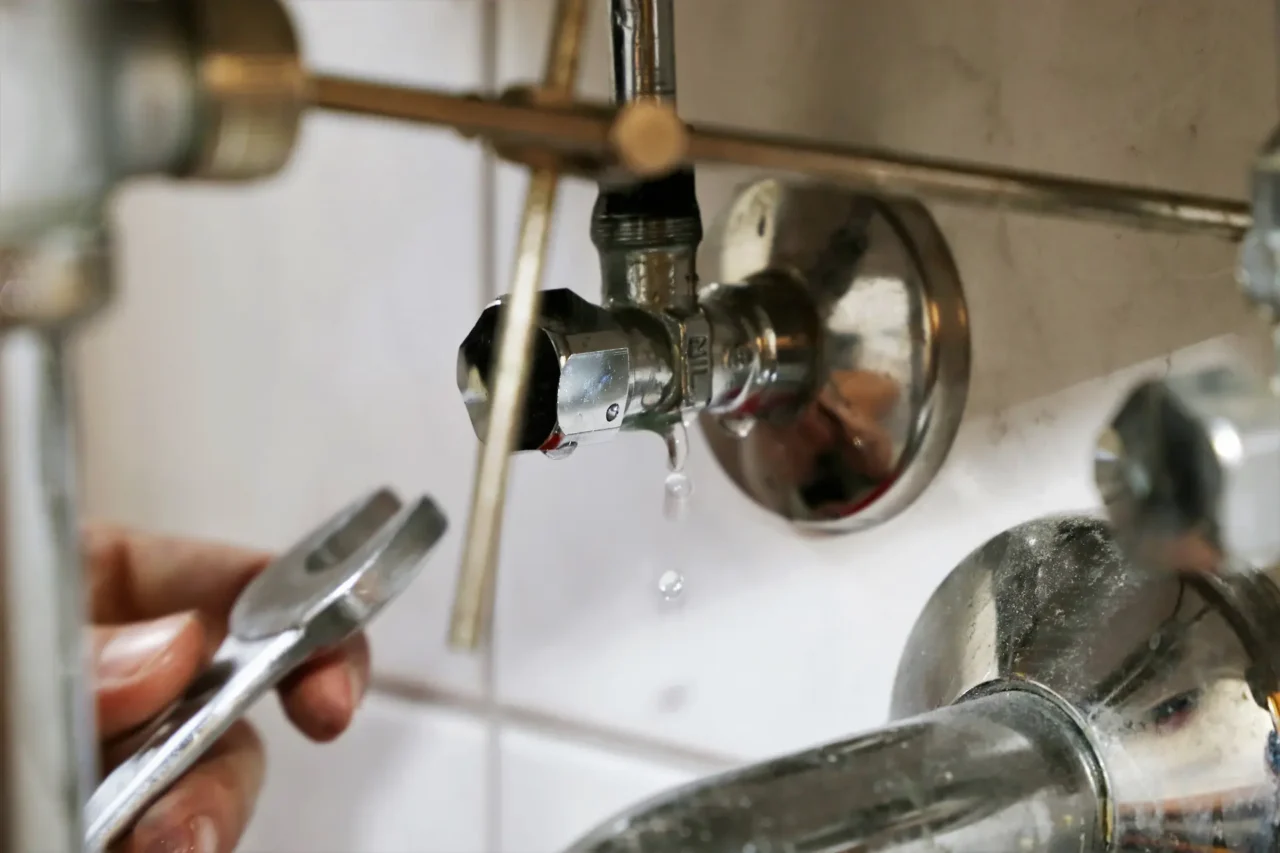 Hand with wrench near dripping chrome valve on tiled wall.